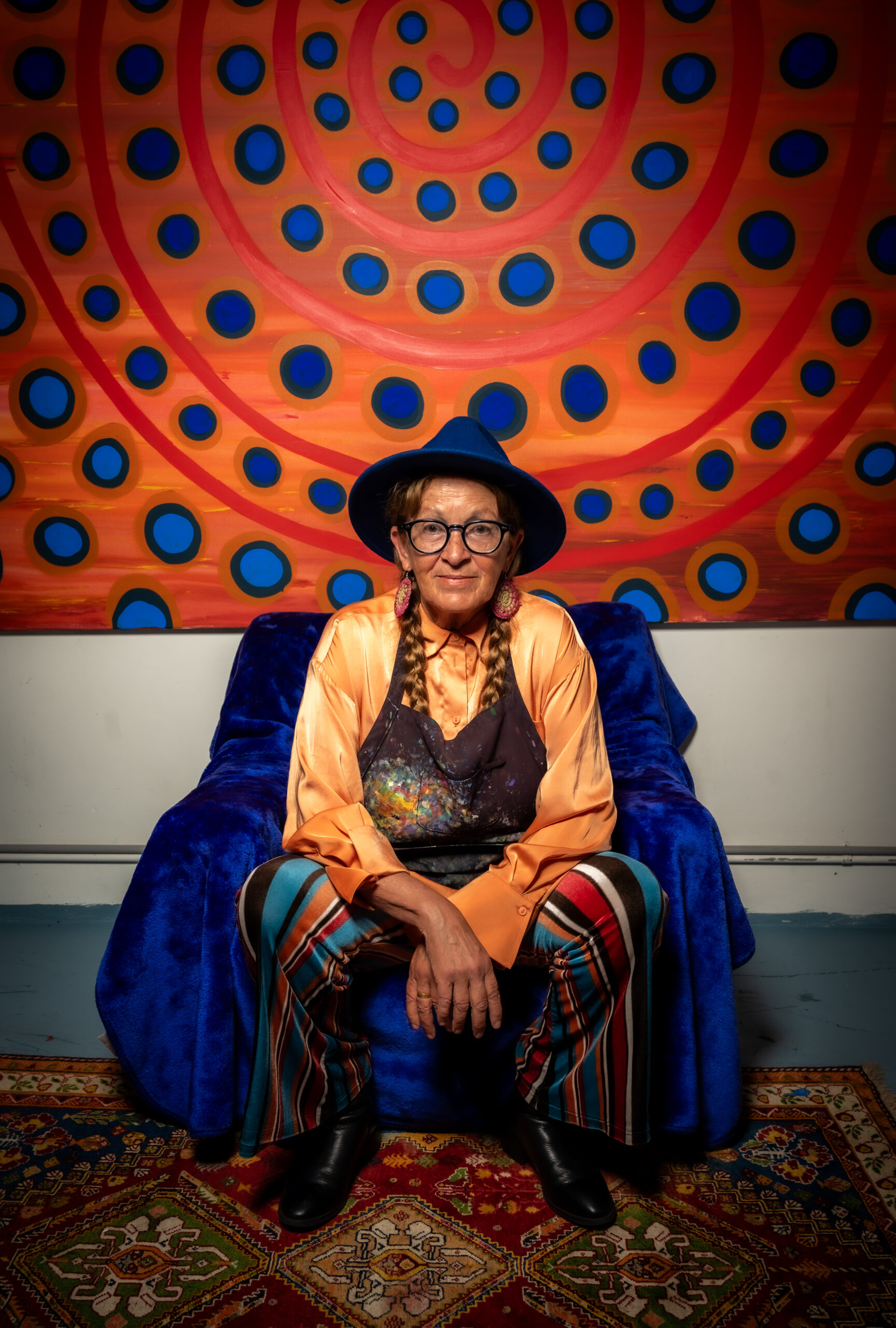 Portrait of an older woman in a blue hat and orange blouse, seated on a blue chair in front of a bright orange wall with blue circles and red spirals.