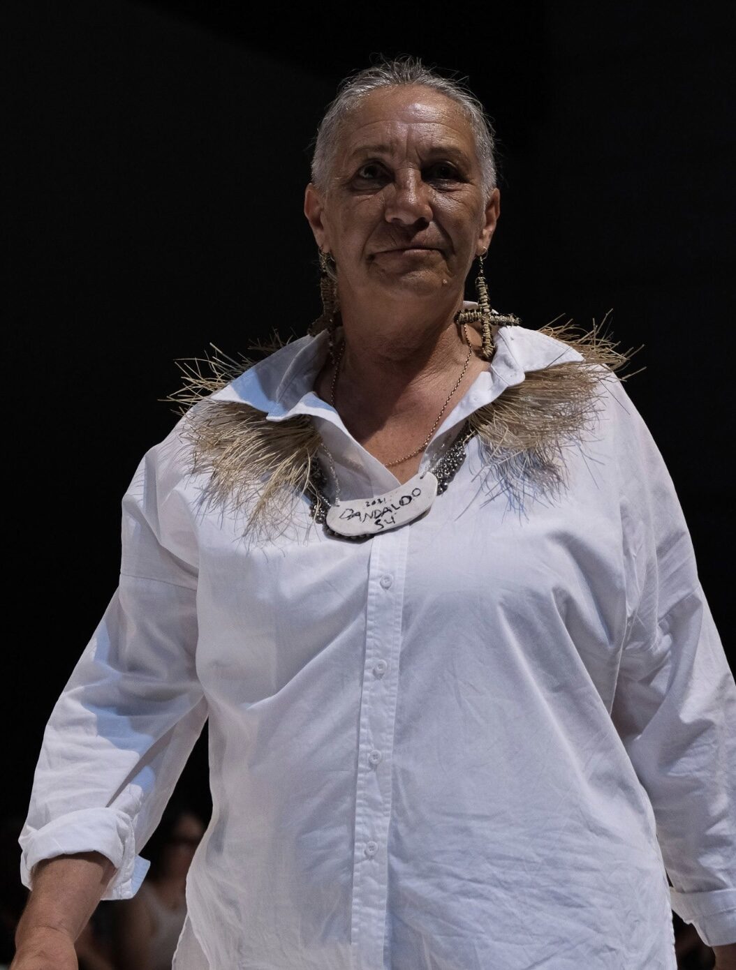 Performer on stage wearing a white button-down shirt with a spiky straw-like collar, large earrings, and a handwritten name tag.