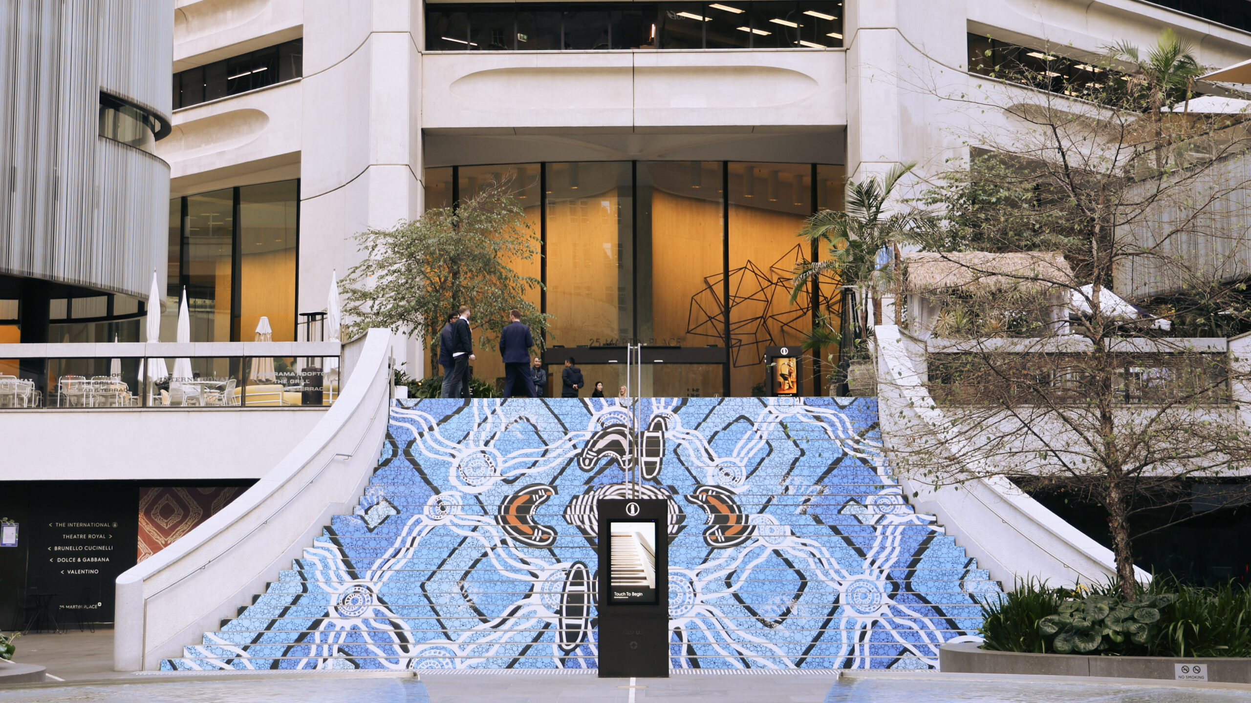 Outdoor stairway covered in a vivid blue geometric mural leading up to a modern glass-front building; several people stand on the upper level.